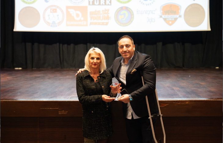 Sahnenin önünde iki kişi yan yana duruyor; ikisi de kameraya dönük ve ellerinde birlikte tuttukları bir ödül bulunuyor. Arkada çeşitli kurum logolarının yer aldığı büyük bir projeksiyon perdesi var. Ortam resmî, tören havasında; sahne ışıkları ödül anının önemini vurguluyor.