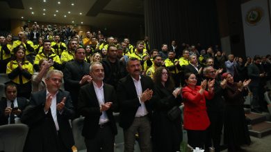 Bir konferans salonunda, ön sıralarda resmi giyimli katılımcılar, arka sıralarda sarı montlu kalabalık bir grup ayakta alkışlıyor; atmosfer coşkulu ve tören havasında.