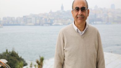 Görselde, arka planda deniz ve şehir manzarasıyla bir adam poz veriyor. Adam, açık renk bir kazak giymiş ve gülümsüyor. Arkadaki şehir silueti net bir şekilde görülüyor. Adamın üzerinde gözlük var ve hava biraz puslu ya da sisli görünüyor.