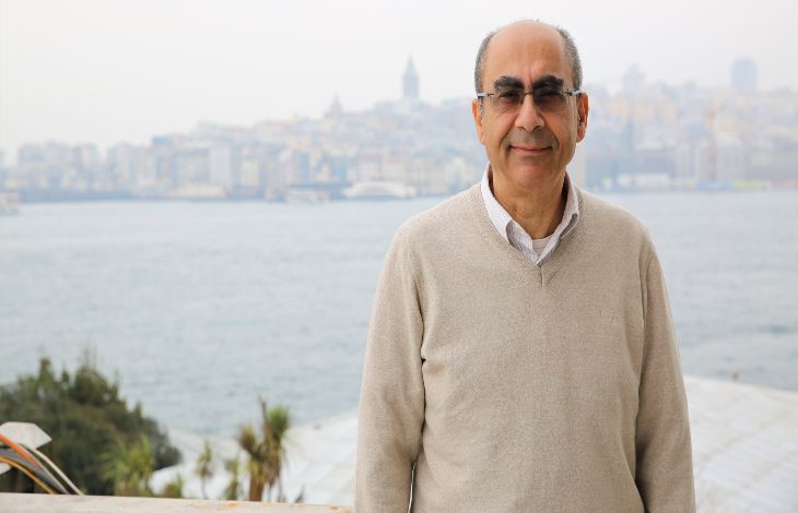 Görselde, arka planda deniz ve şehir manzarasıyla bir adam poz veriyor. Adam, açık renk bir kazak giymiş ve gülümsüyor. Arkadaki şehir silueti net bir şekilde görülüyor. Adamın üzerinde gözlük var ve hava biraz puslu ya da sisli görünüyor.