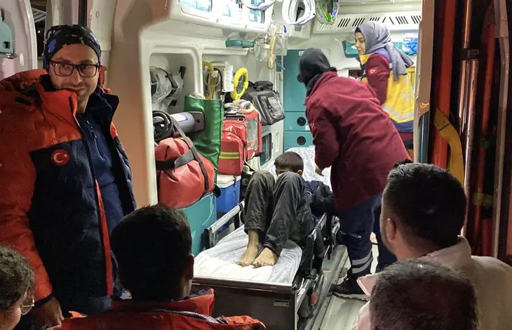 Görselde, bir ambulansın içinde sedyeye yatırılmış bir çocuğa sağlık ekipleri müdahale ediyor. Etrafında ambulans personeli ve görevliler bulunuyor; ortam acil müdahale durumunu yansıtıyor.