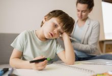 Görselde, bir çocuk masada resim çizerken yorgun veya dalgın bir ifadeyle başını eline dayamış halde görülüyor. Arkada bir yetişkin oturup dikkatle bir şey inceliyor; ortam sakin ve iç mekân.