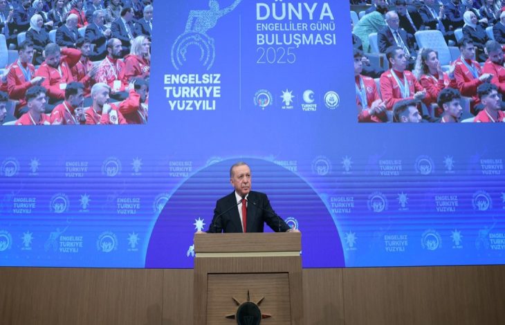 Cumhurbaşkanı Recep Tayyip Erdoğan, büyük bir sahnenin önünde kürsüde konuşma yapıyor, arka planda Dünya Engelliler Günü Buluşması 2025 yazılı mavi bir ekran ve etkinliğe katılan kalabalık bir izleyici grubu görülmektedir.