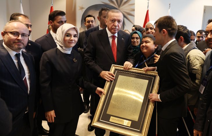 Görselde kalabalık bir grup insan bir arada duruyor. Ortada takım elbiseli bir erkek, büyük boyutlu çerçeveli bir tabloyu iki yanındaki kişilerle birlikte tutuyor. Çerçevenin içinde altın tonlarında bir metin yer alıyor. Sol tarafta başörtülü bir kadın, sağ tarafta ise genç bir erkek bulunuyor. Arkada farklı yaş ve görünümde birçok kişi görüntüye dahil olmuş. Ortam resmi ve tören havasında, arka planda bayraklar görülüyor.