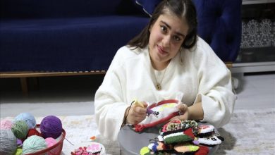 Görselde, ev ortamında yerde oturan genç bir kadın görülüyor. Elinde nakış kasnağıyla panç nakışı yapıyor; etrafında renkli ip yumakları ve tamamlanmış el emeği ürünler yer alıyor. Sahne, üretken ve sakin bir çalışma anını yansıtıyor.