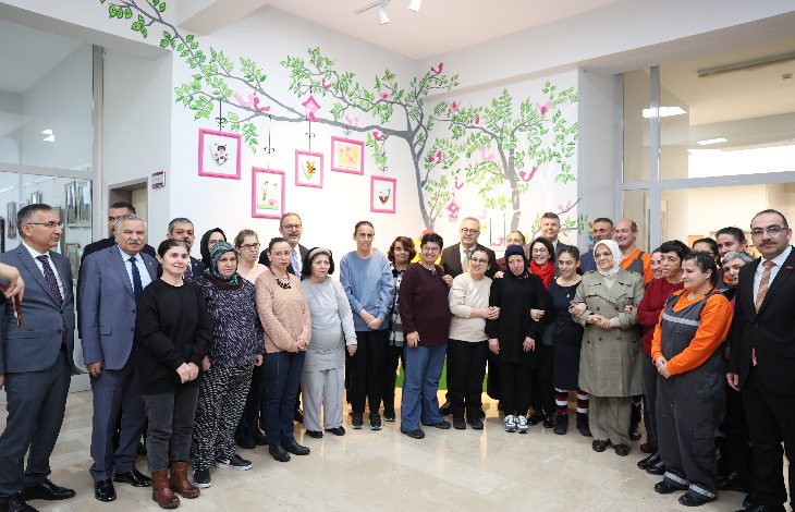 Bir bakım ve rehabilitasyon merkezinde, duvarları ağaç ve kuş figürleriyle süslenmiş aydınlık bir alanda, farklı yaş ve profillerden kalabalık bir grubun yan yana durarak toplu hatıra fotoğrafı çektirdiği görülüyor. Katılımcılar samimi ve resmi karışımı bir ortamda kameraya bakıyor.