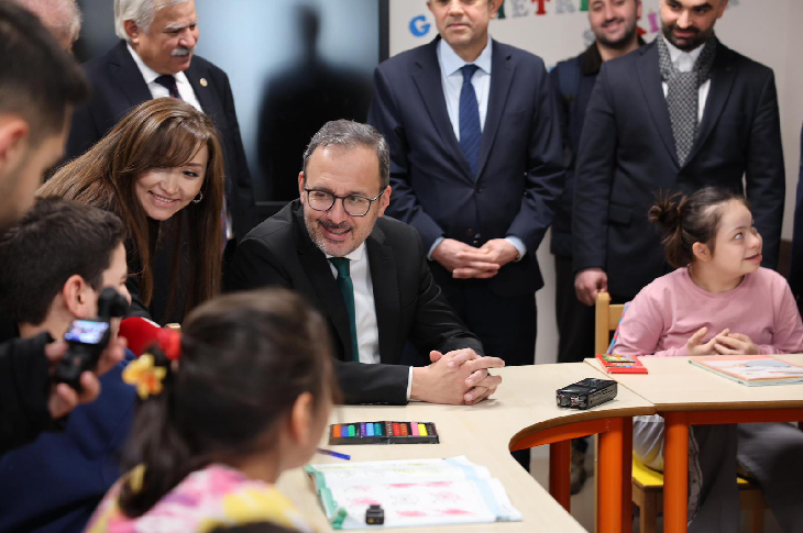 Bir sınıf ortamında, masanın etrafında toplanmış çocuklarla sohbet eden takım elbiseli bir yetkili öne çıkıyor; çocuklar resim ve boya kalemleriyle meşgulken etraflarında öğretmenler ve katılımcılar dikkatle izliyor.