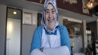Başörtülü bir kadın, mavi üst ve beyaz önlükle bir kafe ortamında kollarını kavuşturmuş şekilde kameraya gülümsüyor; arka planda kafenin tezgâhı ve iç mekânı görülüyor.