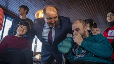 Bir erkek, tekerlekli sandalyede oturan iki engelli bireyin yanına eğilerek onlarla samimi bir şekilde konuşuyor; ortam kalabalık ve sıcak, arka planda başka insanlar da görülüyor.