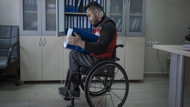 Görselde, bir ofis ortamında tekerlekli sandalyede oturan bir erkek görülüyor. Kucağında dosyalar tutuyor ve dikkatle evrakları inceliyor. Arka planda dolaplar ve klasörler yer alırken, sahne çalışma hayatına aktif katılımı ve odaklanmış bir iş anını yansıtıyor.