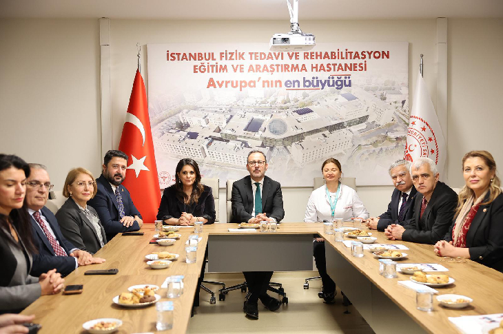 Uzun bir toplantı masasının etrafında toplanmış bir grup yönetici ve yetkili görülüyor. Arkada Türk bayrağı ve Sağlık Bakanlığı amblemi yer alırken, duvarda “İstanbul Fizik Tedavi ve Rehabilitasyon Eğitim ve Araştırma Hastanesi – Avrupa’nın en büyüğü” yazılı pano bulunuyor. Resim, resmi bir ziyaret ya da değerlendirme toplantısı havası taşıyor.