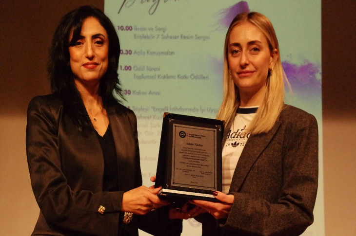 İki kadın sahnede yan yana durarak bir plaket tutuyor. Arkada etkinlik programının yer aldığı bir sunum ekranı görünüyor. Her iki kadın da ciddi ve gururlu bir ifadeyle kameraya bakıyor; ortam bir ödül töreni atmosferinde.