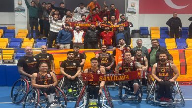 Kapalı bir spor salonunda, Galatasaray Fuzul tekerlekli sandalye basketbol takımı sahada dizilmiş halde poz verirken arkalarında sarı-kırmızı atkılar taşıyan coşkulu taraftarlar, tribün koltukları ve Türk bayrağıyla birlikte birlik ve destek havası görülüyor.