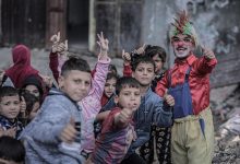 Yıpranmış bir sokak ortamında, renkli kostümlü bir palyaço gülümseyerek başparmağını kaldırırken etrafındaki çocuklar da neşeyle kameraya doğru aynı hareketi yapıyor.