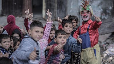 Yıpranmış bir sokak ortamında, renkli kostümlü bir palyaço gülümseyerek başparmağını kaldırırken etrafındaki çocuklar da neşeyle kameraya doğru aynı hareketi yapıyor.