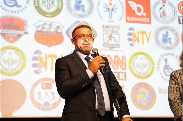 Görselde, takım elbiseli bir erkek sahnede mikrofonla konuşuyor. Arkadaki ekranda çeşitli kurum ve dernek logoları yer alıyor.