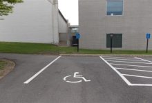 Bir binanın önünde, zeminde tekerlekli sandalye simgesiyle işaretlenmiş engelli park yeri görülüyor. Park alanı boş, çizgiler net; arkada bina girişi ve yönlendirme tabelaları yer alıyor.
