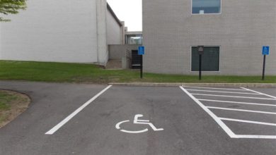 Bir binanın önünde, zeminde tekerlekli sandalye simgesiyle işaretlenmiş engelli park yeri görülüyor. Park alanı boş, çizgiler net; arkada bina girişi ve yönlendirme tabelaları yer alıyor.