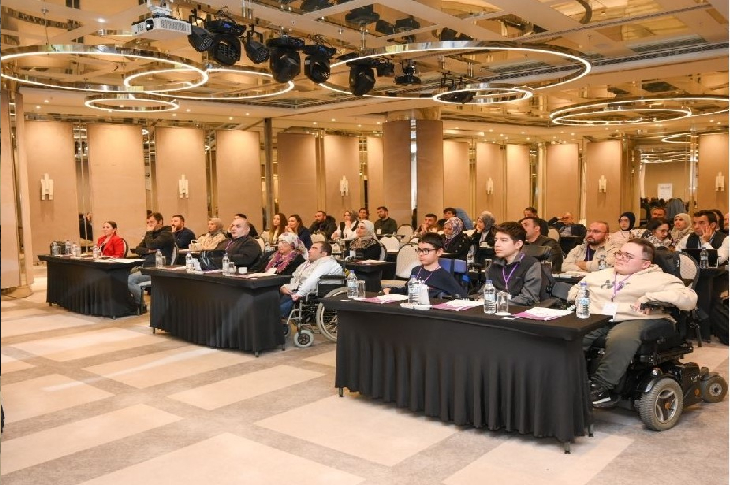 Bir konferans salonunda, tekerlekli sandalye kullanan bireylerin de yer aldığı katılımcıların masalarda oturarak SMA hastalığına yönelik bilgilendirici bir semineri dikkatle dinlediği görülüyor.