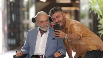 Işık alan bir mekânda, tekerlekli sandalyede oturan Haluk Bilginer ile yanındaki Feyyaz Yiğit'in gülümseyerek birlikte telefonla selfie çektiği görülüyor.