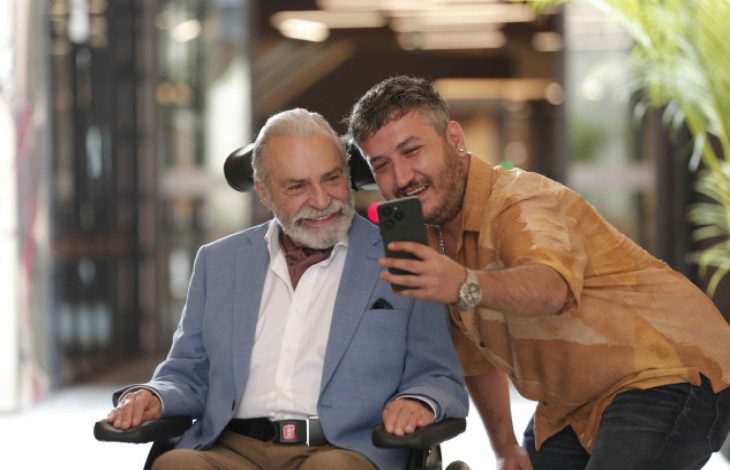 Işık alan bir mekânda, tekerlekli sandalyede oturan Haluk Bilginer ile yanındaki Feyyaz Yiğit'in gülümseyerek birlikte telefonla selfie çektiği görülüyor.