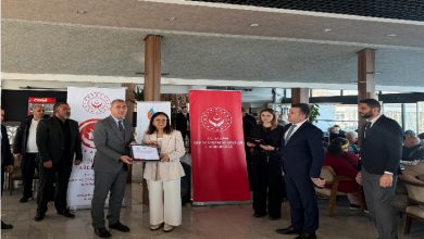 Fotoğrafta bir tören anı görülüyor. Ortada, bir kadın ile bir erkek birlikte bir sertifika tutarak kameraya poz veriyor. Arkalarında Aile ve Sosyal Hizmetler Bakanlığı logolu iki büyük pankart bulunuyor. Sağda ve solda takım elbiseli yetkililer ile davetliler ayakta duruyor. Mekân ferah, gün ışığı alan bir salon ve arkada oturan kalabalık bir topluluk dikkat çekiyor.