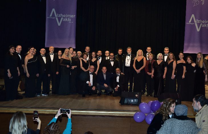 Bir sahnede, şık siyah kıyafetler giymiş kalabalık bir koro ve katılımcılar toplu halde poz veriyor; arkada Alzheimer Vakfı afişleri, önde ise izleyiciler fotoğraf çekiyor.