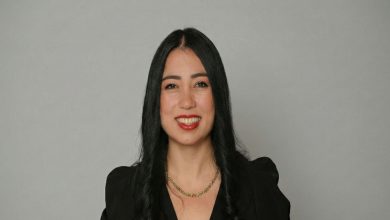 Düz, sade bir arka plan önünde duran, siyah saçlı ve siyah bir kıyafet giymiş bir kadın portresi görülüyor. Kadın kameraya gülümseyerek bakıyor; fotoğraf profesyonel ve resmi bir tanıtım çekimi havası taşıyor.