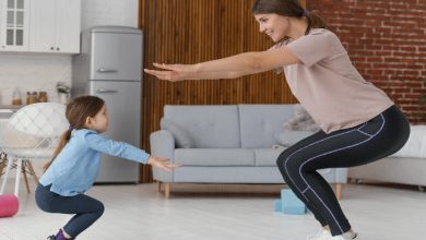 Görselde, ev ortamında bir yetişkin kadın ile küçük bir kız çocuğu birlikte çömelme egzersizi yapıyor. İkisi de birbirine bakarak aynı hareketi tekrar ediyor ve eğlenceli bir an paylaşıyor.