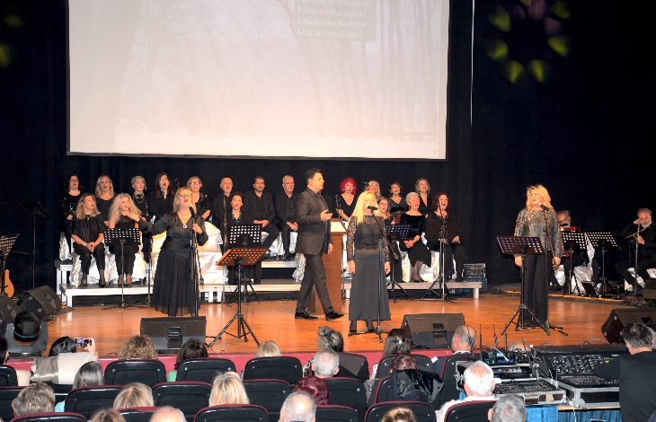 Bir konser salonunda sahnede koro ve solistlerin yer aldığı, arkada müzisyenlerin bulunduğu kalabalık bir müzik dinletisi görülüyor. Ön planda seyirciler otururken, sahnede toplu performans ve canlı bir etkinlik atmosferi dikkat çekiyor.