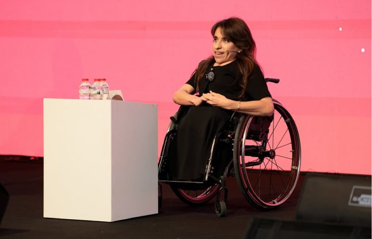 Görselde, tekerlekli sandalye kullanan bir kadın sahnede konuşma yaparken görülüyor. Önünde beyaz bir kürsü ve üzerinde su şişeleri bulunuyor. Arka plan canlı pembe renkte ve kadın profesyonel bir ortamda, mikrofonuyla birlikte izleyicilere hitap ediyor gibi görünüyor.