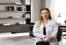 Görselde, modern bir ofis ortamında beyaz önlük giymiş bir kadın oturuyor ve kameraya doğru gülümseyerek poz veriyor.