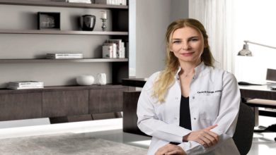 Görselde, modern bir ofis ortamında beyaz önlük giymiş bir kadın oturuyor ve kameraya doğru gülümseyerek poz veriyor.