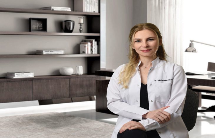 Görselde, modern bir ofis ortamında beyaz önlük giymiş bir kadın oturuyor ve kameraya doğru gülümseyerek poz veriyor.