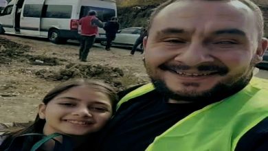 Gülümseyerek kameraya bakan bir yetişkin erkek ve yanında genç bir kız görülüyor. Erkek fosforlu bir yelek giymiş. Arkada, çamurlu bir yol kenarında beyaz bir minibüs ve birkaç kişi yer alıyor.