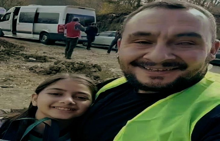 Gülümseyerek kameraya bakan bir yetişkin erkek ve yanında genç bir kız görülüyor. Erkek fosforlu bir yelek giymiş. Arkada, çamurlu bir yol kenarında beyaz bir minibüs ve birkaç kişi yer alıyor.