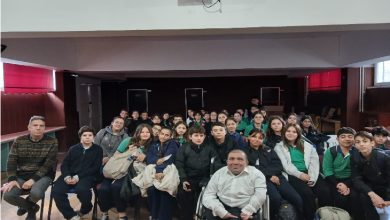 Bir konferans salonunda, kalabalık bir öğrenci grubu toplu fotoğraf için kameraya bakıyor. Ön sırada öğretmenler ve tekerlekli sandalyedeki bir konuşmacı yer alıyor. Arka sıraları dolduran öğrenciler meraklı ve neşeli bir ifadeyle poz veriyor. Salonun ışığı yumuşak, ortam sıcak ve samimi.