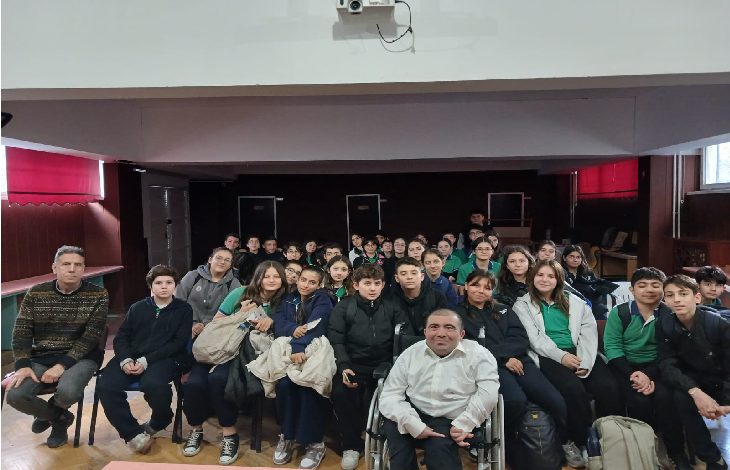 Bir konferans salonunda, kalabalık bir öğrenci grubu toplu fotoğraf için kameraya bakıyor. Ön sırada öğretmenler ve tekerlekli sandalyedeki bir konuşmacı yer alıyor. Arka sıraları dolduran öğrenciler meraklı ve neşeli bir ifadeyle poz veriyor. Salonun ışığı yumuşak, ortam sıcak ve samimi.