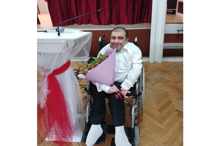 Fotoğrafta, tekerlekli sandalyede oturan bir kişi elinde büyük ve renkli bir çiçek buketiyle kameraya gülümsüyor. Yanında kırmızı-beyaz tüllerle süslenmiş bir kürsü var. Arka planda bordo bir sahne perdesi görülüyor. Ortam sıcak, teşekkür ve takdir duygusunun havada hissedildiği bir anı yansıtıyor.