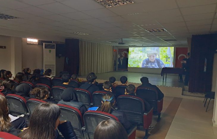 Bir konferans salonunda sıralara oturmuş öğrenciler, perdede yansıtılan bir belgeseli dikkatle izliyor. Işıklar kısık, sahnede sunum alanı ve Türk bayrağı var. Ortam, farkındalık ve empati odaklı bir etkinliğin sessiz ama yoğun havasını taşıyor.