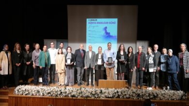 Bir sahnede yan yana dizilmiş kalabalık bir grup topluca poz veriyor. Arkadaki büyük ekranda “3 Aralık Dünya Engelliler Günü” başlıklı program afişi görünüyor. Önde çiçeklerle süslenmiş bir platform var. Bazı katılımcılar plaket veya ödül benzeri şeyler tutuyor; ortam resmi, akademik ve tören havasında.