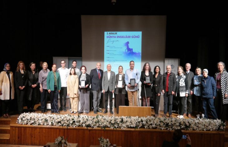 Bir sahnede yan yana dizilmiş kalabalık bir grup topluca poz veriyor. Arkadaki büyük ekranda “3 Aralık Dünya Engelliler Günü” başlıklı program afişi görünüyor. Önde çiçeklerle süslenmiş bir platform var. Bazı katılımcılar plaket veya ödül benzeri şeyler tutuyor; ortam resmi, akademik ve tören havasında.