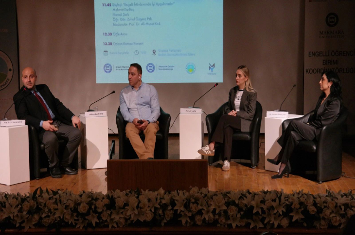 Bir panel sahnesi var: Dört kişi yarım daire şeklinde yerleştirilmiş sandalyelerde oturuyor. Her birinin önünde isimlikler bulunuyor. Arkada, etkinliğin program akışını gösteren büyük bir sunum ekranı yer alıyor. Panelistler ciddi ve odaklanmış bir şekilde konuşmaya hazır görünürken, ortam akademik ve resmi bir tartışma atmosferi taşıyor.