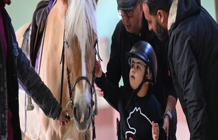 Bir kapalı alanda, kask takmış küçük bir çocuk bir atın yanında duruyor. Yanında bulunan iki yetişkin, çocuğu dikkatle yönlendirirken biri atın başını tutuyor. Sahne sakin ve odaklı; terapi amaçlı bir etkinlikte güven ve temas ön planda.