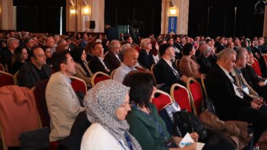 Fotoğrafta büyük bir salonun içi görülüyor. Kırmızı sandalyelere sıralar hâlinde oturmuş çok sayıda katılımcı, sahne yönüne doğru dikkatle dinliyor. Çoğu takım elbiseli ya da resmi giyimli. Ortam bir konferans ya da sempozyum havasında; salon aydınlık, duvarlarda büyük panolar ve kameralar dikkat çekiyor. Salonda yoğun bir katılım olduğu hemen fark ediliyor.