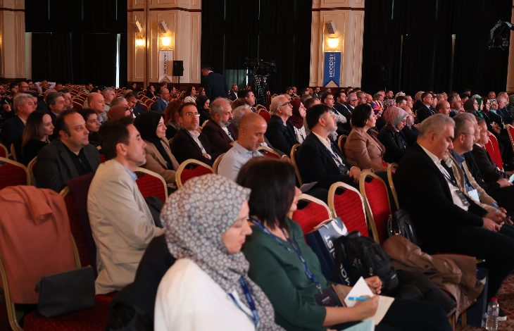 Fotoğrafta büyük bir salonun içi görülüyor. Kırmızı sandalyelere sıralar hâlinde oturmuş çok sayıda katılımcı, sahne yönüne doğru dikkatle dinliyor. Çoğu takım elbiseli ya da resmi giyimli. Ortam bir konferans ya da sempozyum havasında; salon aydınlık, duvarlarda büyük panolar ve kameralar dikkat çekiyor. Salonda yoğun bir katılım olduğu hemen fark ediliyor.