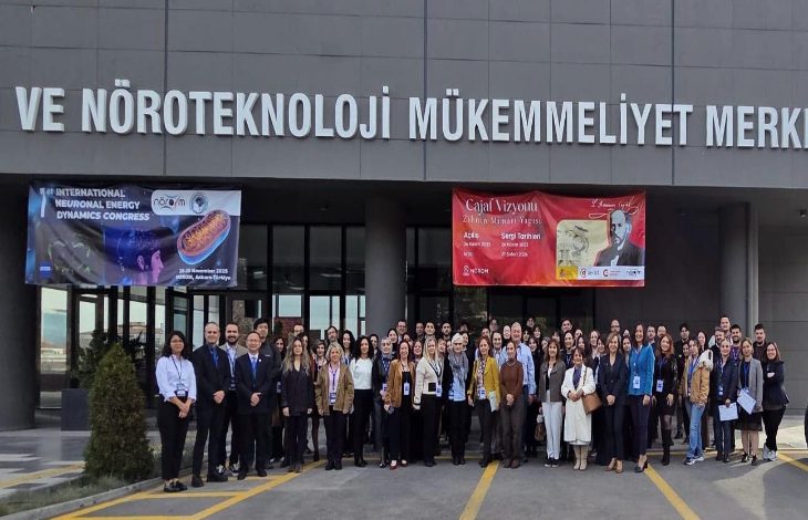 Binanın girişinde geniş bir grup, omuz omuza dizilmiş şekilde toplu fotoğraf veriyor. Üstte “Nöroteknoloji Mükemmeliyet Merkezi” tabelası ve iki büyük kongre afişi görülüyor. Katılımcılar resmi ama enerjik bir duruşla, bilimsel bir etkinliğin açılış anını yansıtıyor.