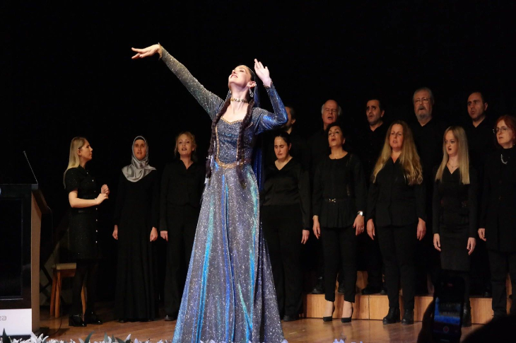 Görselde sahnede yapılan bir gösteri yer alıyor. Ön planda, ışıltılı uzun bir elbise giyen bir kadın sahne ortasında kollarını yukarı doğru açmış etkileyici bir performans sergiliyor. Arkada ise tamamı siyah kıyafetli bir koro sıralanmış şekilde duruyor ve performansa eşlik ediyor. Sahne ışıkları sanatçıyı öne çıkarırken, arka planda koro üyeleri ciddi ve odaklanmış bir duruş sergiliyor. Genel atmosfer, sanatsal ve duygusal bir sahne gösterisini yansıtıyor.