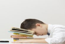 Fotoğrafta, küçük bir çocuk bir masa başında yorgun şekilde uyuklarken görülüyor. Başını, üst üste dizilmiş renkli kitapların üzerine koymuş. Üzerinde beyaz gömlek ve kravat var. Arka plan sade ve boş; bu da çocuğun yorgunluğunu ve ders yükünü daha belirgin kılıyor.