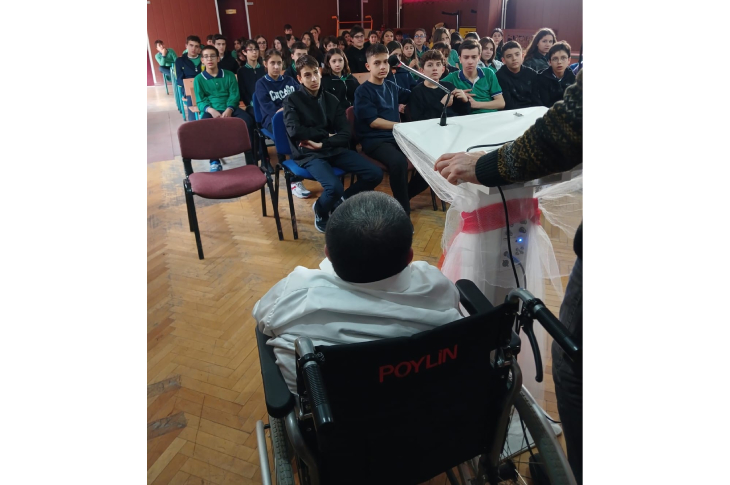Fotoğrafta bir konuşma anının arka tarafından sahneye bakıyoruz. Ön planda tekerlekli sandalyede oturan bir kişi kürsüye yönelmiş durumda. Kürsünün başında mikrofonu tutan konuşmacının kolu görülüyor. Karşı tarafta, sıralar dolusu öğrenci dikkatle dinliyor; yüzlerindeki merak, yoğunlaşma hâli net biçimde seçiliyor. Salonun zemini ahşap, ortam ciddi ama aynı zamanda içten bir etkileşime sahne oluyor.